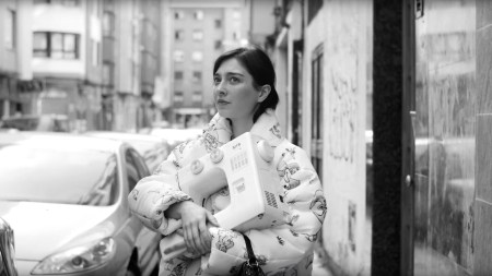 A black-and-white film still showing a dark-haried Argentian girl holding a sewing machine and wearing a puff coat.