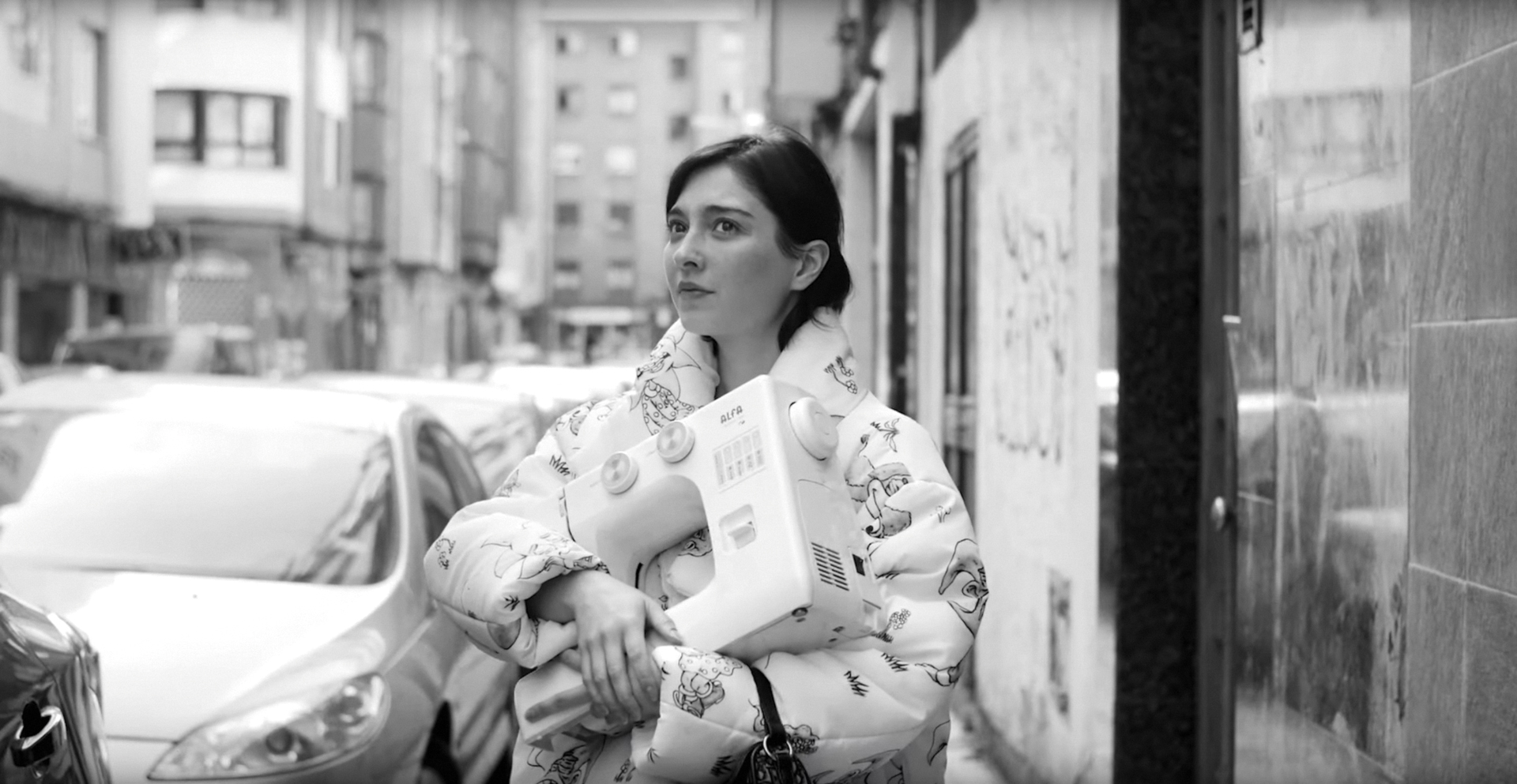 A black-and-white film still showing a dark-haried Argentian girl holding a sewing machine and wearing a puff coat.