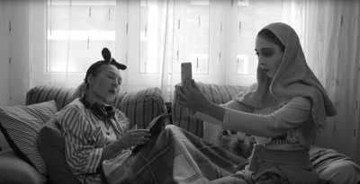 Two women lounge on a couch. One is wearing a towel and a face mask while taking a selfie. The other is looking at a screen and has a pair of headphones around her neck.