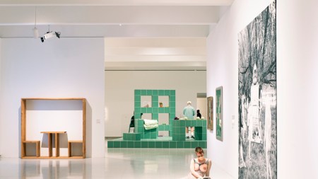 Within a museum space, a tapestry hangs on a wall and extends onto the floor, where two performers lounge. A green tiled platform and a sculpture of wooden chairs appears in the background.