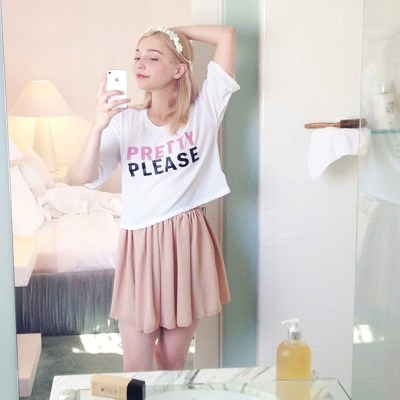 A blonde girl in a hotel room is wearing a shirt that says pretty please, and a pink skirt.
