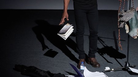A photograph of a man's legs and one arm as he drops a book. There are other papers on the floor and at the far right a mic stand with feathers and a horse-head mask hanging from it.