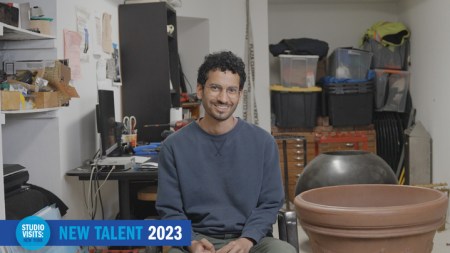 A brown man with short curly dark hair and glasses sits on a chair, smiling looking at the camera.