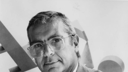 An older man in a suit and ties poses with his arms crossed.