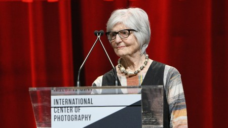 A woman standing at a podium before a red curtain.