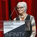 A woman standing at a podium before a red curtain.
