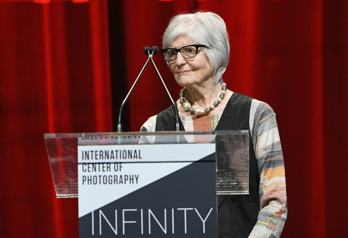 A woman standing at a podium before a red curtain.