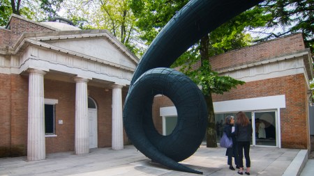 People standing outside a large curving sculpture outside a columned pavilion.