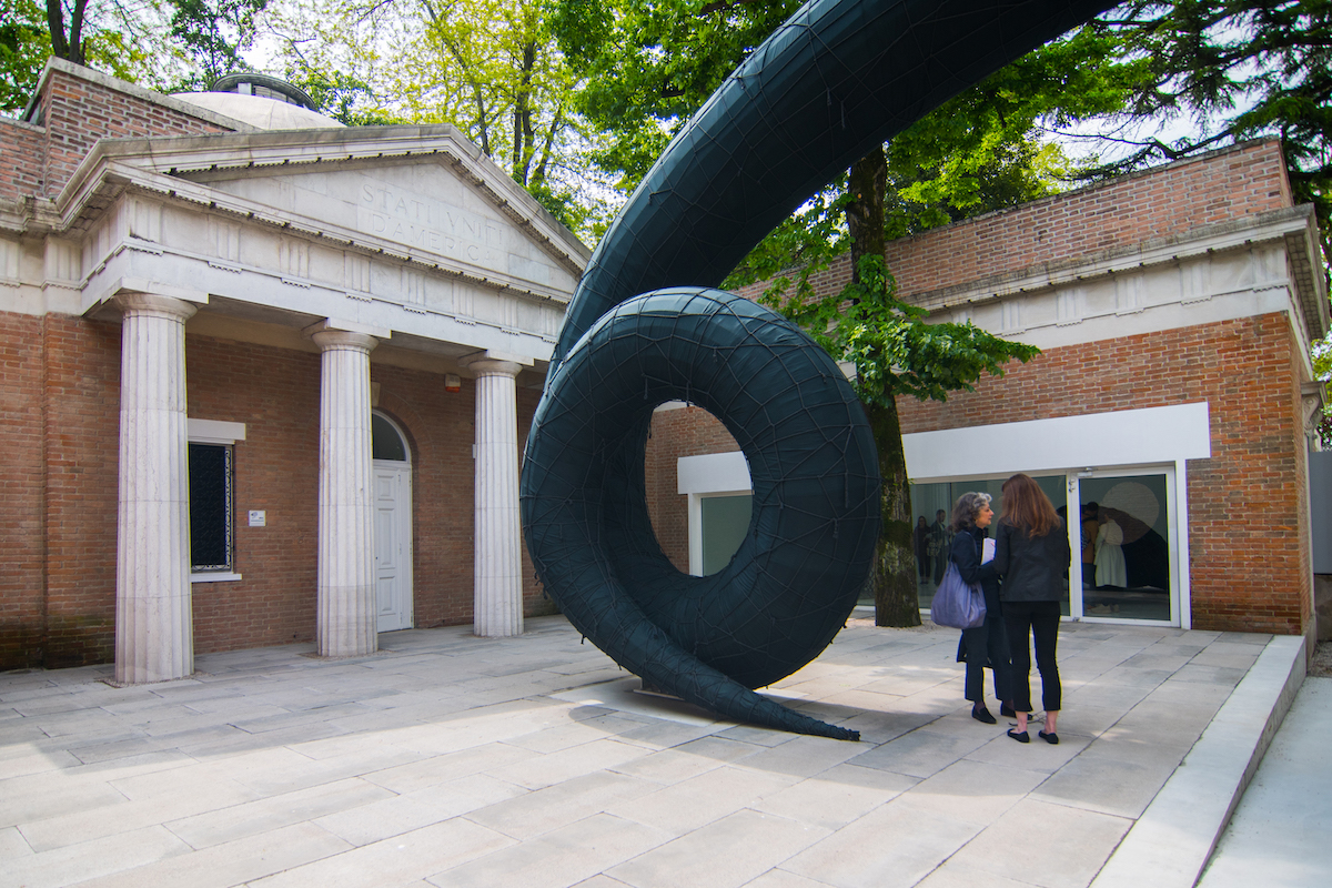People standing outside a large curving sculpture outside a columned pavilion.