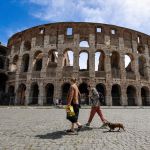 Two women walk a dog past an ancient building.