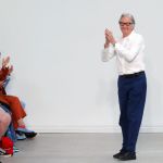 An older gentleman in a white collared shirt, blue pants, and black shoes applauds in front of a white wall as a small audience of people does too.