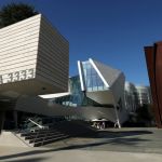 Exterior of a newly built museum showing three distinct structures. At left is rectangular shape made of light wood that juts over a small staircase. At center is a silver-ish building with angles and glass. At right is a Richard Serra sculpture made of brown steel.