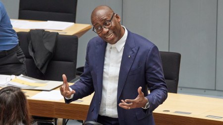 A Black man in a suit stands before a low table with his briefcase on it. He holds his hands up and tilts his head to the side.