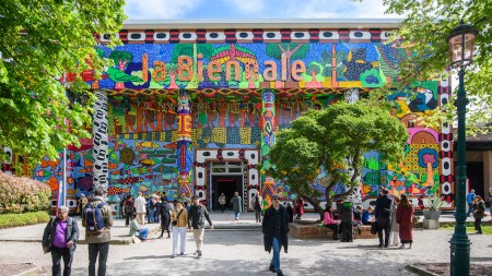 People walking in and out of a treed courtyard in front of a brightly painted building with columns. On its roof, there is the phrase 'la Biennale.'