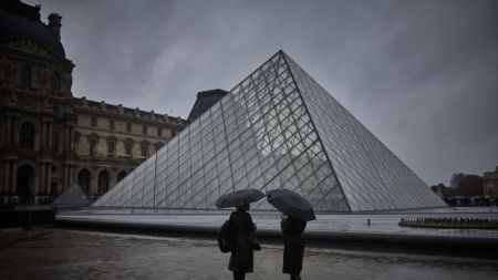 Two people shelter under an umbrella during a rainy day next to a glass pyramid structure.