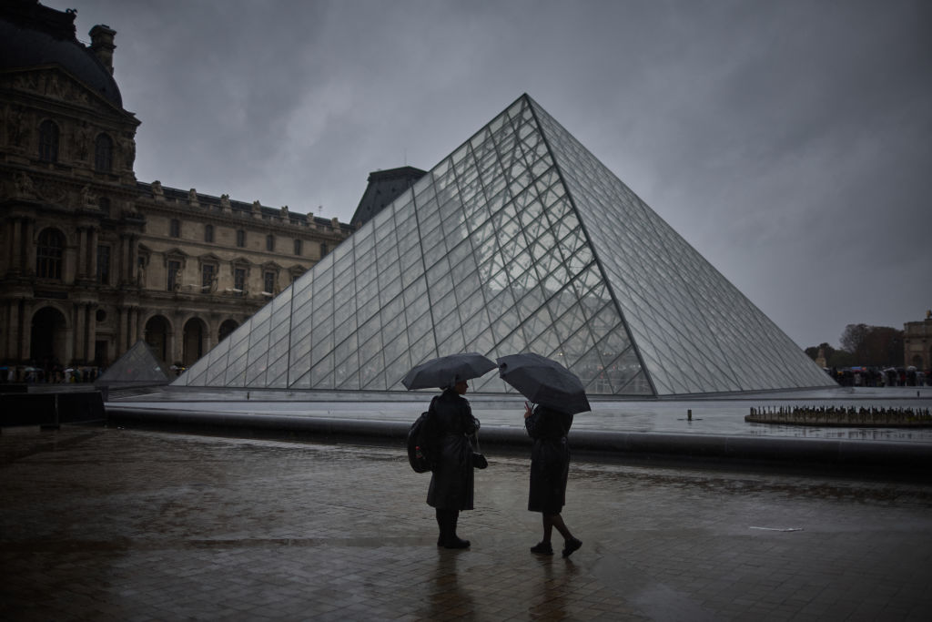 Two people shelter under an umbrella during a rainy day next to a glass pyramid structure.