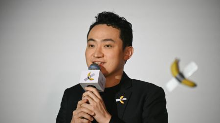 A man stands in front of a banana duct-taped to a wall holding a microphone.