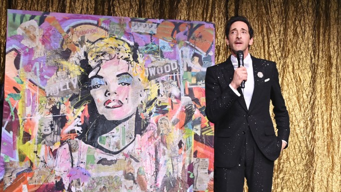 A man in a tuxedo beside a painting of Marilyn Monroe collaged with various materials.