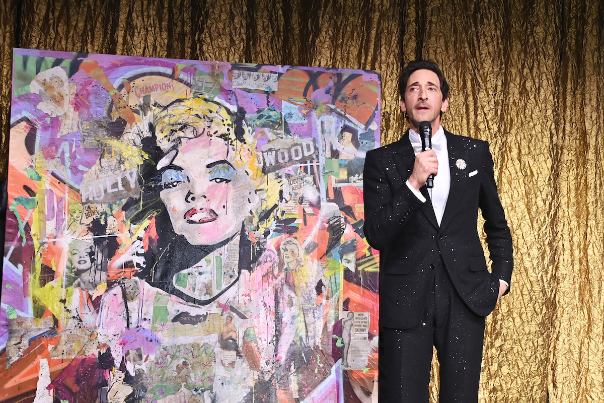 A man in a tuxedo beside a painting of Marilyn Monroe collaged with various materials.