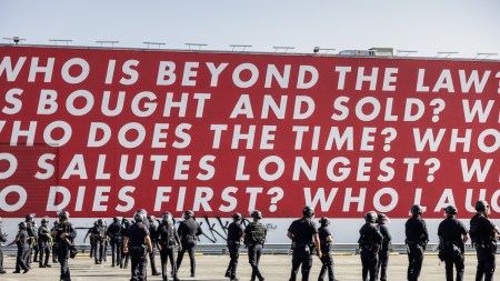 A group of soldiers beneath a red mural with text that begins 'WHO IS BEYOND THE LAW?'