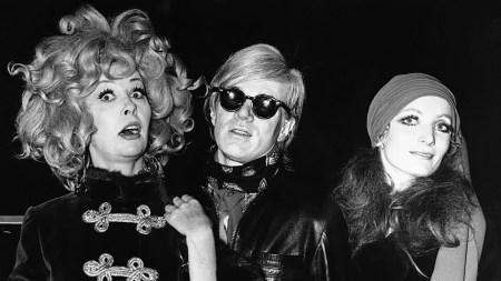 A black-and-white photo showing Andy Warhol and two glamorous women, all of them dressed in black.