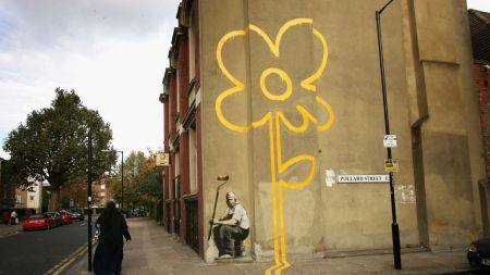 A woman walks past a some a painting of a yellow flower on a wall on a street.