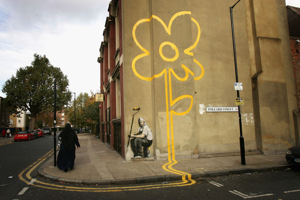 A woman walks past a some a painting of a yellow flower on a wall on a street.
