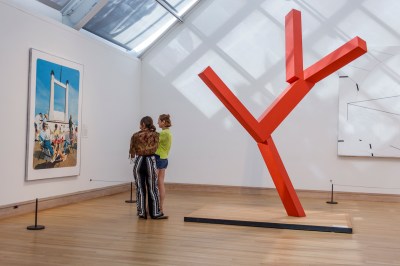 Two women staring at a painting in a sunlit gallery beside a tall orange-colored sculpture resembling a stick figure.