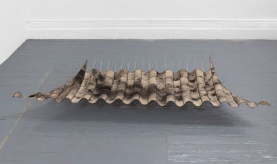A sculpture of what appears to be charred paper floats above a gray wood floor. 