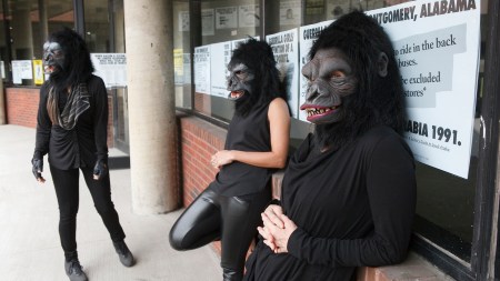 Three women dressed all in black with gorilla masks over their heads are leaning against a brick wall with a window. The window is covered in posters with big black letters.