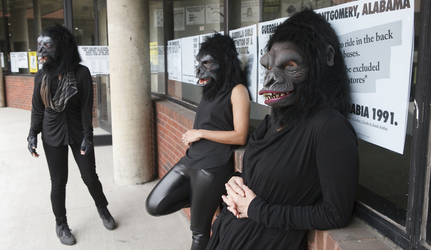 Three women dressed all in black with gorilla masks over their heads are leaning against a brick wall with a window. The window is covered in posters with big black letters.