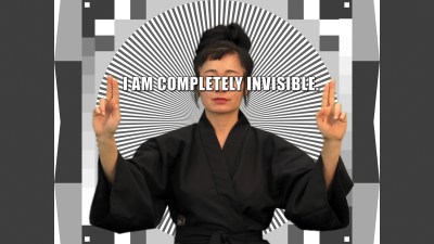 An Asian woman in a black robe holds up both her hands with two fingeres each. In between them are the words I AM COMPLETELY INVISIBLE.