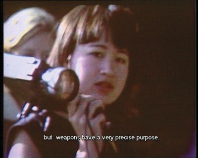 In a LoFi video still, a young Hito Steyerl, an East Asian woman, holds a video camera. A caption says: But weapons have a very precise purpose.