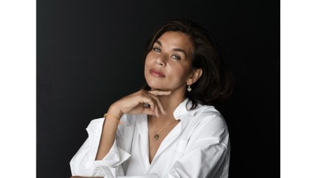 Portrait of a Black woman wearing a white shirt. She is pictured against an all black backdrop and her hands rest on a pedestal.