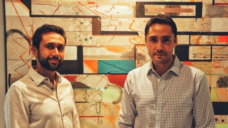 Two Brown men in button-down shirts stand before an abstract painting.