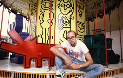 Keith Haring, a white bearded man with glases, sits in front of a carousel based on his iconic designs, colorful cartoony figures with bold outlines.