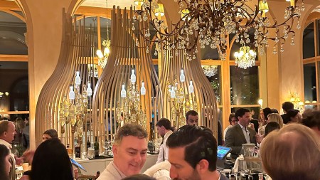 Several people stand around a bar, lit up by gold chandeliers.