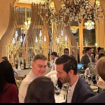 Several people stand around a bar, lit up by gold chandeliers.