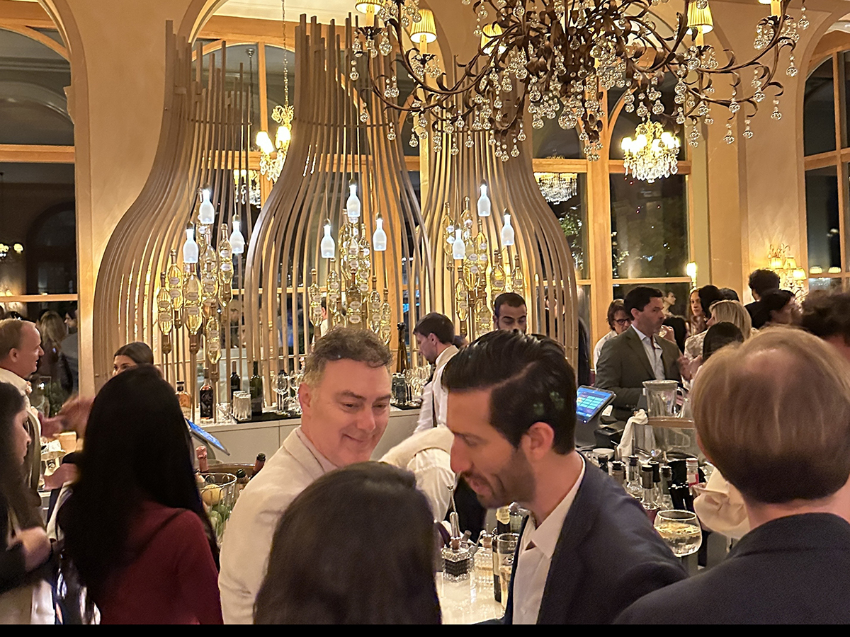 Several people stand around a bar, lit up by gold chandeliers.