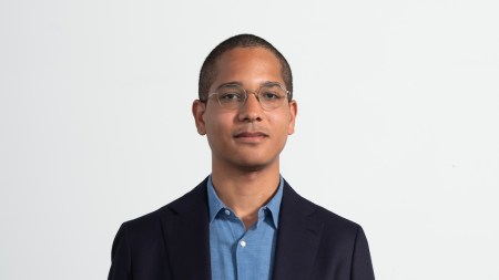 A young man with glasses and short hair in a blue button up shit and dark blue blazer.