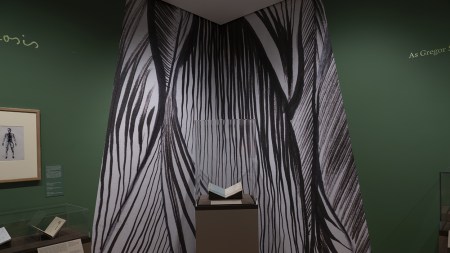 A manuscript on a pedestal in the corner of a green room. A graphic wall drawing is made of black lines and looks like a tunnel or a curatin.
