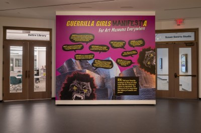A blowup of a poster with text and images of gorilla masks. Yellow and black texts include one saying, “Guerrilla Girls Manifesta for art museums everywhere.” 