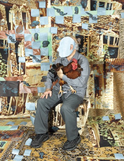 A man holds a rooster in a photo studio. An ornate mustard carpet is beneath him and the wall behind him is collaged in a grid-olike pattern, with drawings of snakes and pictures of old buildings.