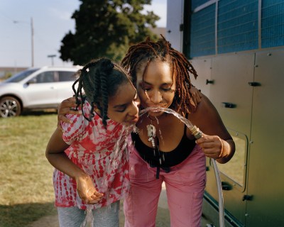 Two Black women, a mother and daughter in Spring clothes, drink from a hose with their arms around each other.