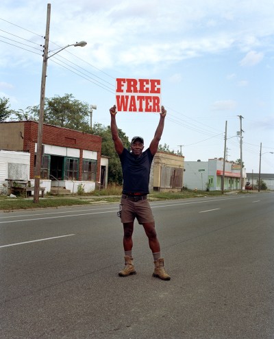 A Black man wearing shorts and hiking boots stands in the middle of Rustbelt road holding a sign that says FREE WATER in red.