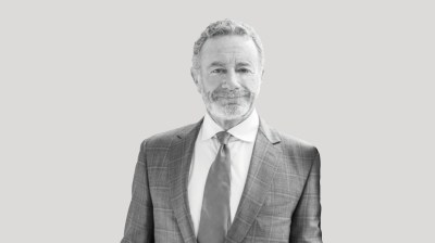 A silhouetted black-and-white portrait of a white man in a suit and tie on a gray background. He has a half smile.
