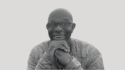 A silhouetted black-and-white portrait of a smiling Black man with glasses on a gray background. His hands are folded at his chin.