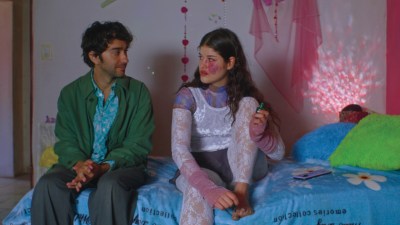 A man and woman sit on a bed in what looks like a teenager's room. The man has his hands folded in his lap as he looks into her eyes; he looks somewhat nervous or uncomfortable. The girl, wearing a lot of purple lace and velvet, is painting her toes purple; she has a large purple and red spot on her left cheek.