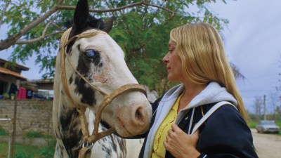 Chloë Sevigny, a white celebrity with long blonde hair, gazes into the eyes of a spotted horse with visible cataracts.