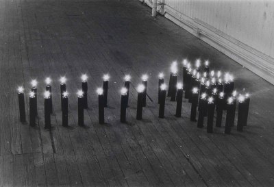 A black-and-white photograph showing a cross-like shape made of lit dark candles.  